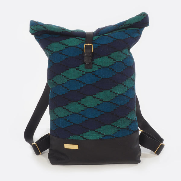 Front view of Battiayo ,Deep Blue Sea' backpack with handwoven traditional dhaka fabric from Nepal and leather. The handwoven fabric of the backpack has three different shades of blue and a bit of black. The dhaka fabric has a geometric pattern and is handwoven by amazing women. The leather is black. The straps and bottom are made from leather. The brass buckles and brass label are also made in Nepal.