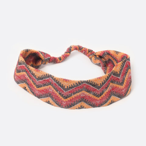 Top view of the Battiayo Sun & Moon Red headband. The handwoven traditional dhaka fabric is red, orange, black and a bit white. The dhaka fabric has a geometric pattern and is handwoven by amazing women. The pattern is a zigzag pattern. The small scrunchy part, which gives the headband the elasticity is shown. The elastic part is covered with the dhaka fabric.
