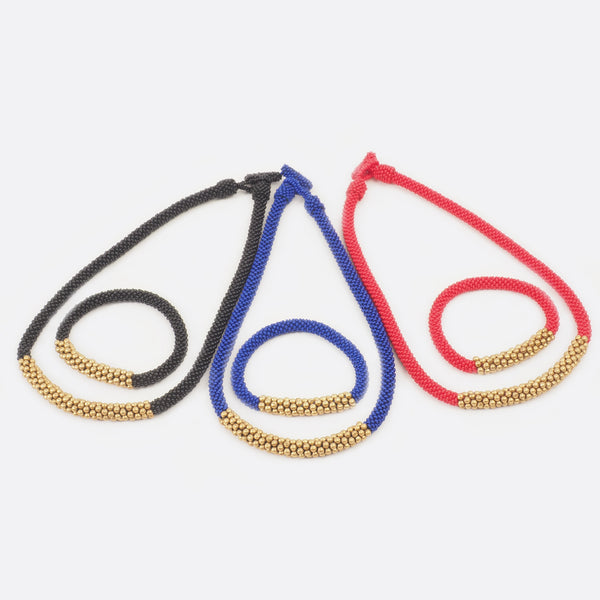 The handmade Battiayo beaded necklace with brass exists in three different colors. The black necklace with the matching bracelet is on the left. The blue necklace with the matching bracelet is in the middle and the red necklace with the matching bracelet is on the left. The background is white.