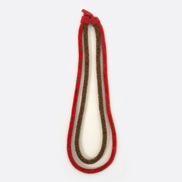 Front view of the Battiayo beaded necklace. It is a double-string necklace. One string is a bit longer than the other string. One string is red and has small golden dots. The glass beads of the other string are all golden. 