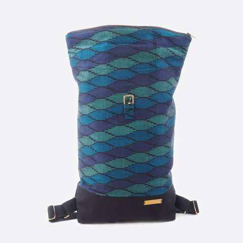 Front view of the Battiayo ,Deep Blue Sea' backpack with handwoven traditional dhaka fabric from Nepal and canvas. The handwoven fabric of the backpack has three different shades of blue and a bit of black. The dhaka fabric has a geometric pattern and is handwoven by amazing women. The canvas is black. The backpack has brass buckles and a brass label. The roll top main compartment is open. Battiayo backpack is visible in its complete length.  The backpack is adaptable in length. 